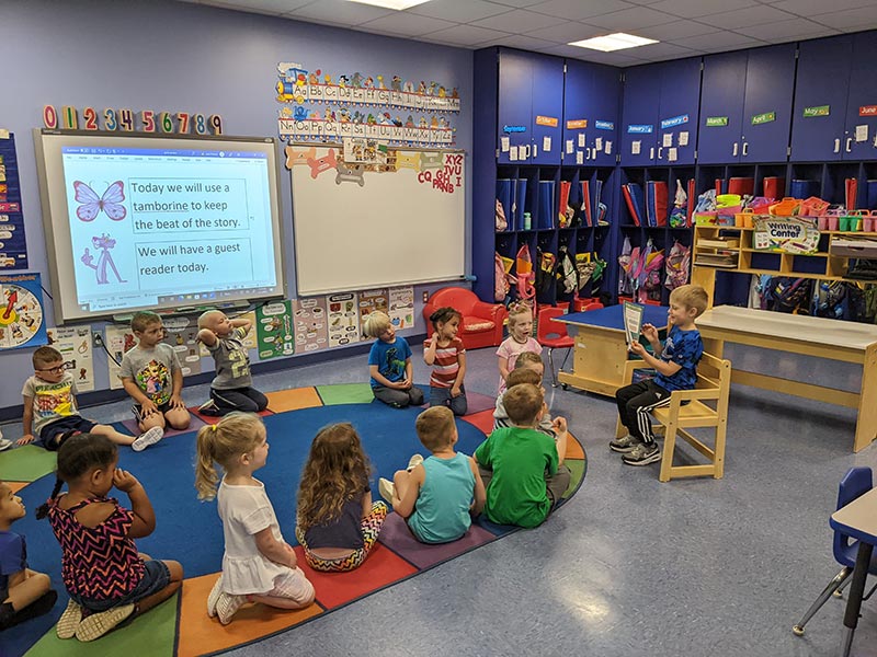 a male student reading to his classmates thta are sitting in a circle on the carpet